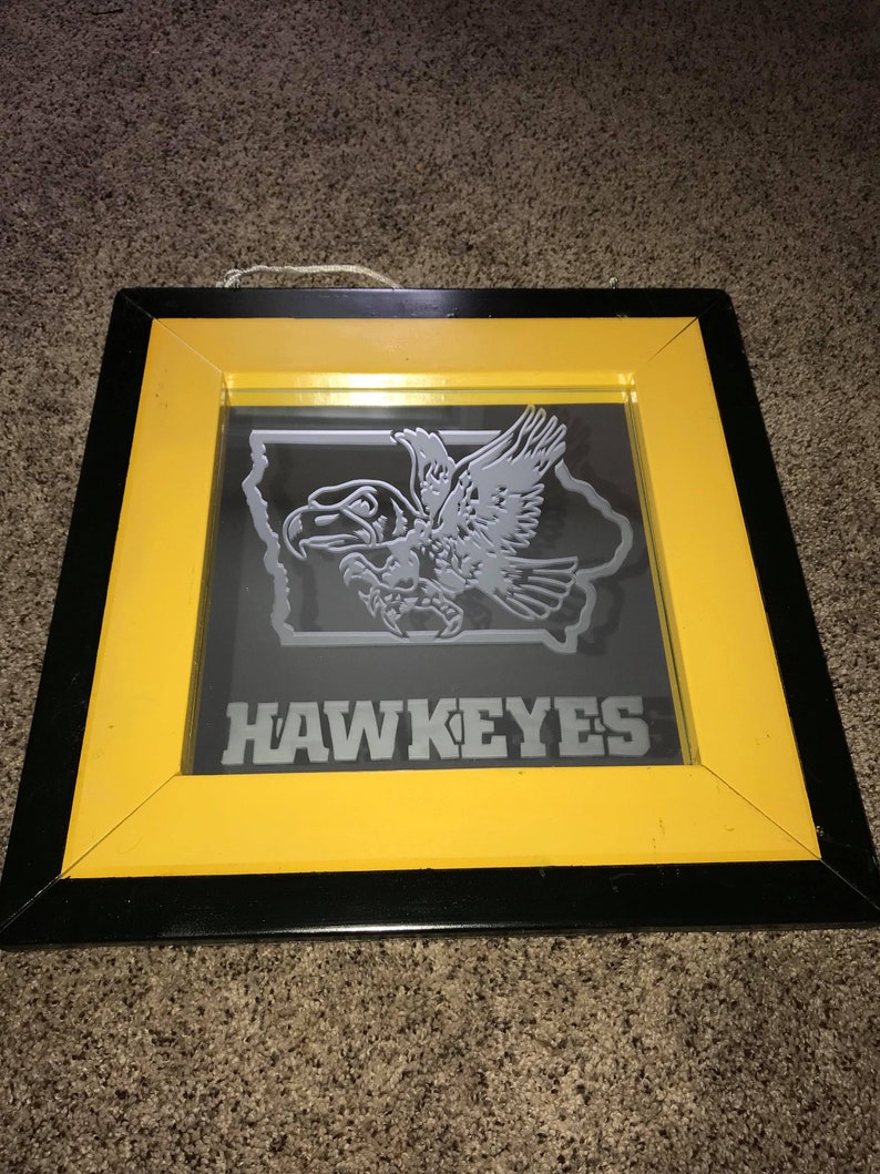 Op de afbeelding: Een ingelijste spiegel met een zwart en geel frame. De spiegel heeft een grijs silhouet van een havik die over de staat Iowa vliegt met het woord "HAWKEYES" eronder.