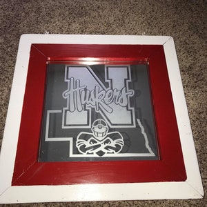 Op de afbeelding: Een ingelijste spiegel met een rood en wit frame. De spiegel heeft een grijze achtergrond met een zilveren silhouet van een voetballer en het woord "Huskers" in zilveren letters.