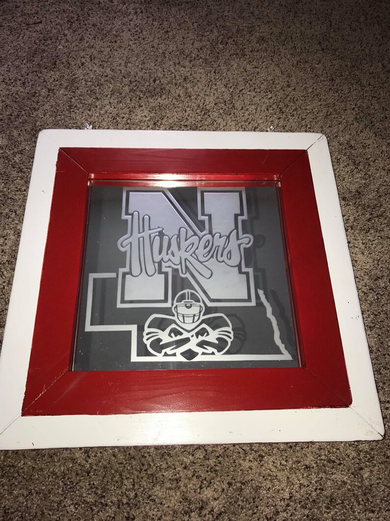 Op de afbeelding: Een ingelijste spiegel met een rood en wit frame. De spiegel heeft een grijze achtergrond met een zilveren silhouet van een voetballer en het woord "Huskers" in zilveren letters.