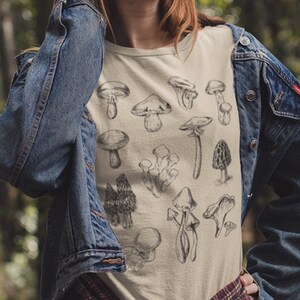 Puede incluir: Una camiseta blanca con una ilustraci&oacute;n en blanco y negro de varios tipos de setas. La camiseta la lleva puesta una persona en un entorno de bosque.