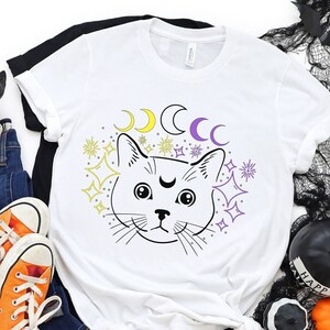 May include: White t-shirt featuring a black and white illustration of a cat's face with a crescent moon above its head. The cat is surrounded by stars and moons in purple, yellow, and white.