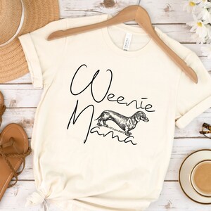 May include: A white t-shirt with the text "Weenie Mama" and a black and white illustration of a dachshund.