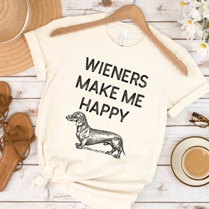 Puede incluir: Una camiseta blanca con una ilustraci&oacute;n de dachshund negra y el texto "Wieners Make Me Happy".