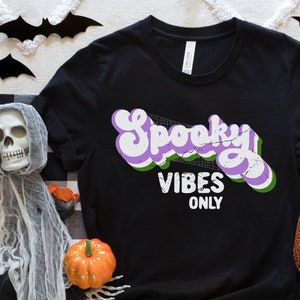 May include: Black t-shirt with a purple and green retro-style graphic that says "Spooky Vibes Only".