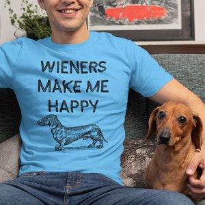 Puede incluir: Un hombre con una camiseta azul claro con el texto "Wieners Make Me Happy" y una ilustraci&oacute;n en blanco y negro de un dachshund. Est&aacute; sentado en un sof&aacute; con un perro dachshund marr&oacute;n.