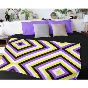 May include: A non-binary pride flag-themed blanket with a geometric diamond pattern in black, yellow, white, and purple. The blanket is displayed on a black bed with purple pillows and neutral-colored throw pillows.