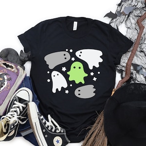 May include: Black t-shirt with a graphic of white and gray ghosts and a green ghost in the center. The ghosts are surrounded by white stars.