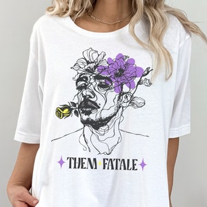 May include: White t-shirt featuring a graphic of a person's face with a flower growing out of their head. The text "Them Fatale" is printed below the graphic.