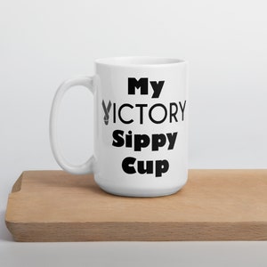 May include: A white ceramic mug with a black handle and the words "My VICTORY Sippy Cup" printed in black. The mug is on a light brown wooden surface.