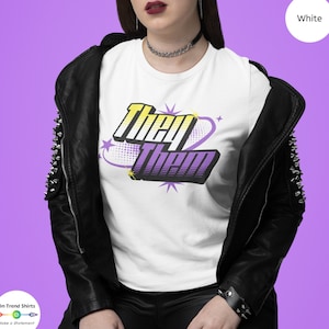 May include: White t-shirt with a purple and yellow graphic that says "They Them" in a comic book style font.