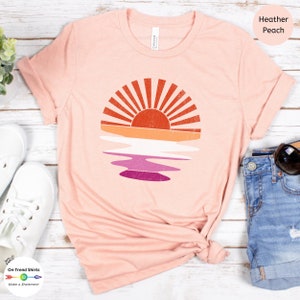May include: A heather peach t-shirt with a vintage-style sunset graphic featuring a rainbow reflection in the water. The graphic is in shades of orange, yellow, pink, and purple.