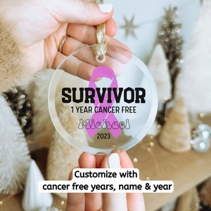 May include: A clear glass ornament with a pink ribbon and the text "SURVIVOR 1 YEAR CANCER FREE Michael 2023".