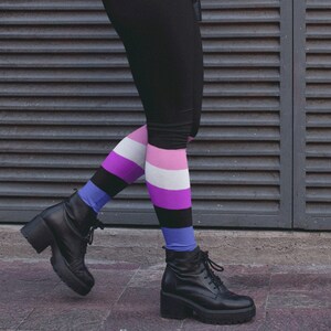 May include: A person wearing black leggings and a black leather jacket with a colourful striped sock on their right leg. The person is standing in front of a grey metal wall.