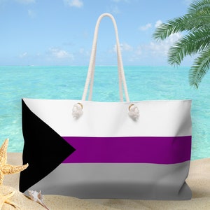 May include: A large tote bag with a white top, a purple stripe, and a grey bottom. The bag features a black triangle on the left side and white rope handles. The background shows a beach scene with a blue ocean and palm trees.