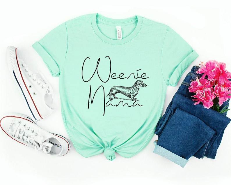 May include: A light green t-shirt with the text "Weenie Mama" in black script. A black and white illustration of a dachshund is above the text.