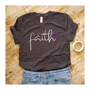 K&ouml;nnte beinhalten: Ein dunkelgraues T-Shirt mit dem Wort "faith" in wei&szlig;er Kursivschrift. Ein lila Band befindet sich neben dem Wort "faith".