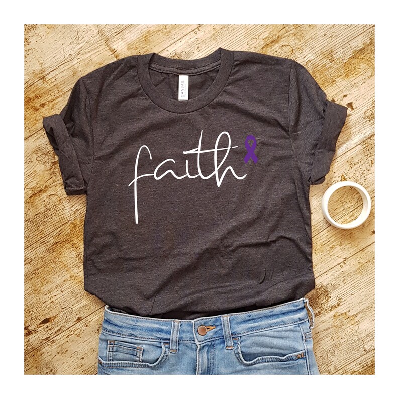 K&ouml;nnte beinhalten: Ein dunkelgraues T-Shirt mit dem Wort "faith" in wei&szlig;er Kursivschrift. Ein lila Band befindet sich neben dem Wort "faith".
