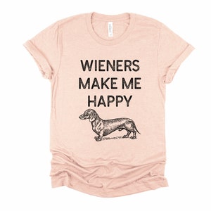 Puede incluir: Una camiseta rosa claro con el texto "Wieners Make Me Happy" y una ilustraci&oacute;n en blanco y negro de un dachshund.