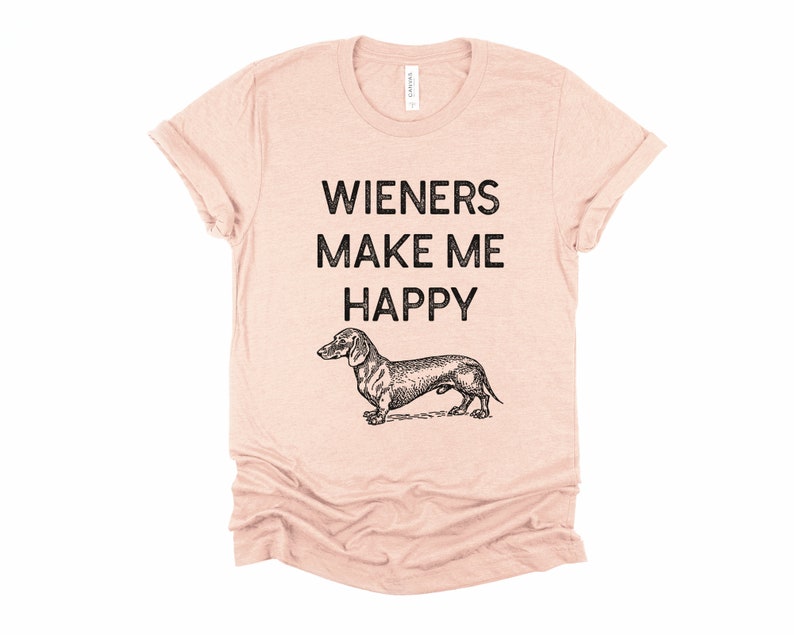 Puede incluir: Una camiseta rosa claro con el texto "Wieners Make Me Happy" y una ilustraci&oacute;n en blanco y negro de un dachshund.