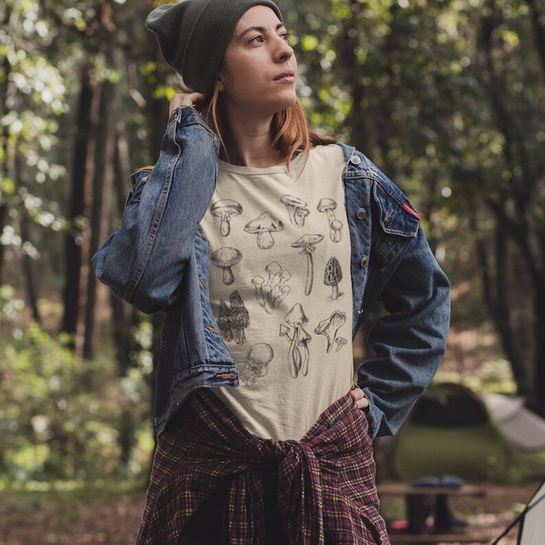 Puede incluir: Una camiseta blanca con una ilustraci&oacute;n en blanco y negro de varios tipos de setas. La camiseta la lleva puesta una persona en un entorno de bosque.