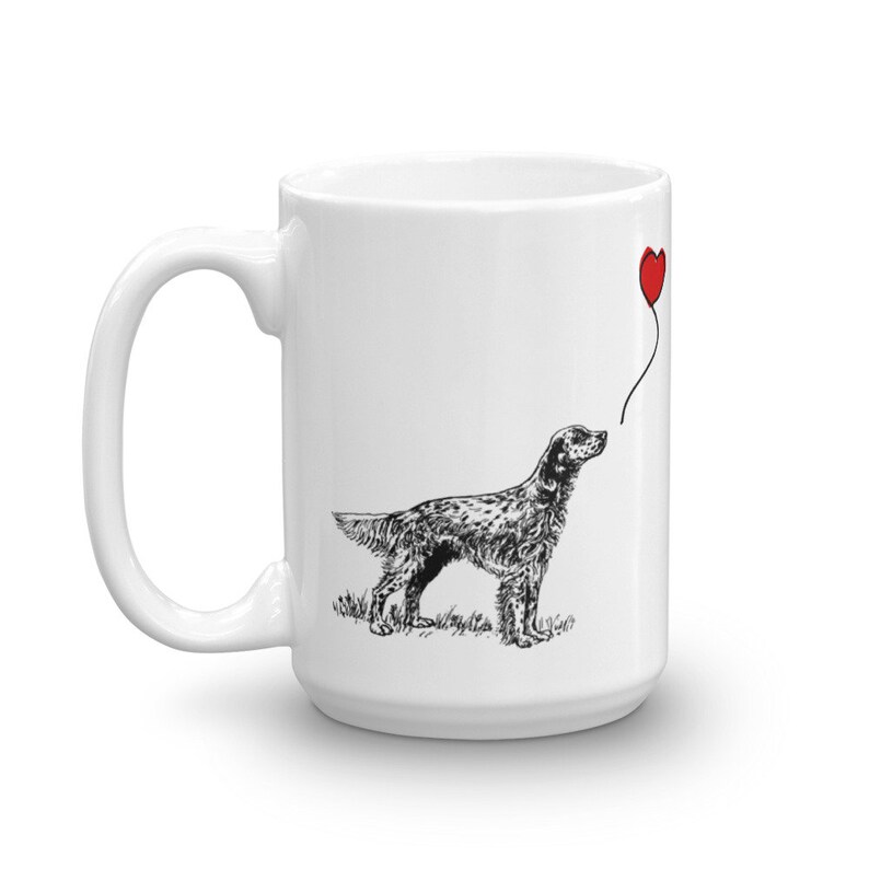 May include: White ceramic mug with a black and white illustration of a dog looking up at a red heart-shaped balloon. The dog is a pointer breed.