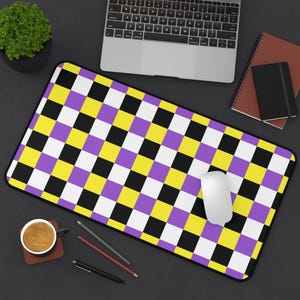 May include: A non-binary pride flag checkered mouse mat with yellow, purple, black and white squares. The mouse mat is on a black surface with a computer mouse, a cup of coffee and pencils.