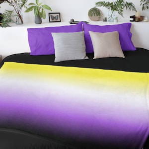 May include: A black and white throw blanket with a yellow, white, and purple ombre design. The blanket is folded on a bed with black sheets and purple and beige pillows.