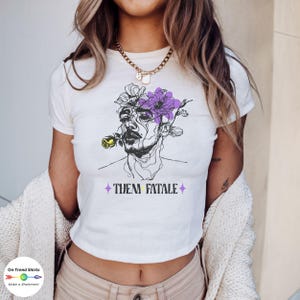 May include: White crop top featuring a black and purple line drawing of a person with a flower growing out of their head. The text "Them Fatale" is printed in yellow below the drawing.