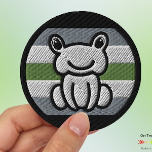 May include: A round embroidered patch featuring a grey frog on a grey, green and black striped background. The patch is approximately 5 cm in diameter.