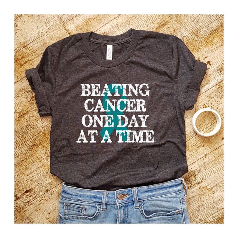 May include: A dark grey t-shirt with the text "BEATING CANCER ONE DAY AT A TIME" in white with a teal ribbon graphic.