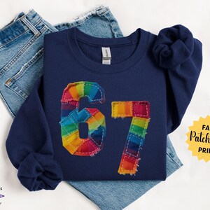 May include: Navy blue sweatshirt with a rainbow patchwork design of the number 67. The sweatshirt is displayed on top of a pair of blue jeans. The words "Faux Patchwork Print" are in a yellow starburst.