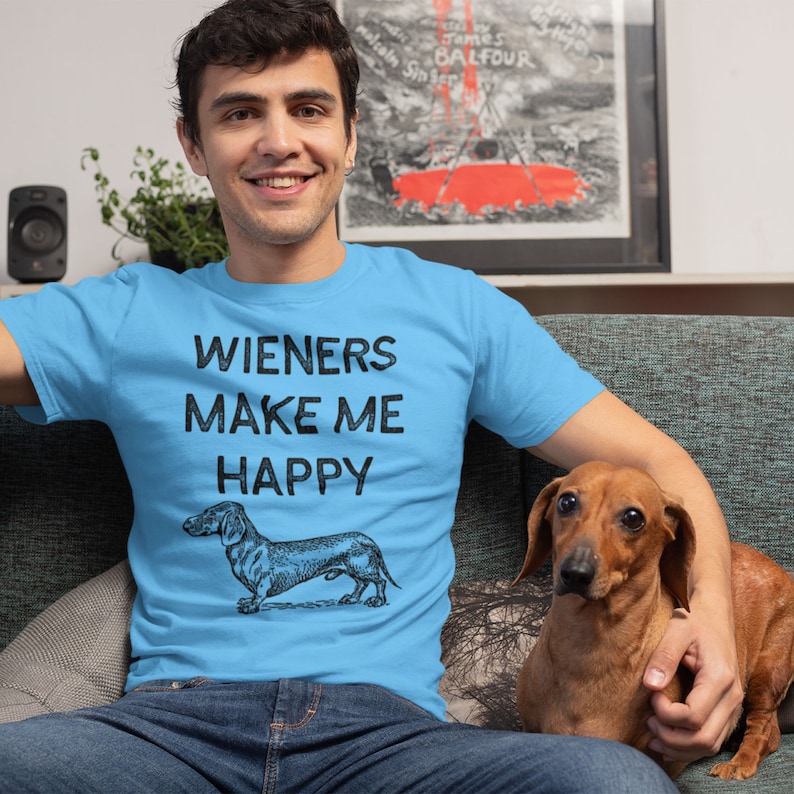 Puede incluir: Un hombre con una camiseta azul claro con el texto "Wieners Make Me Happy" y una ilustraci&oacute;n en blanco y negro de un dachshund. Est&aacute; sentado en un sof&aacute; con un perro dachshund marr&oacute;n.