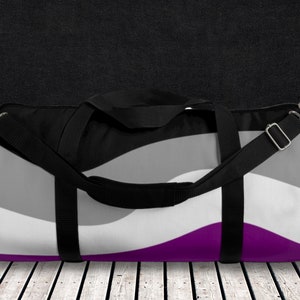 May include: A black duffel bag with a purple, grey, and white striped design. The bag has a black shoulder strap and two black handles.