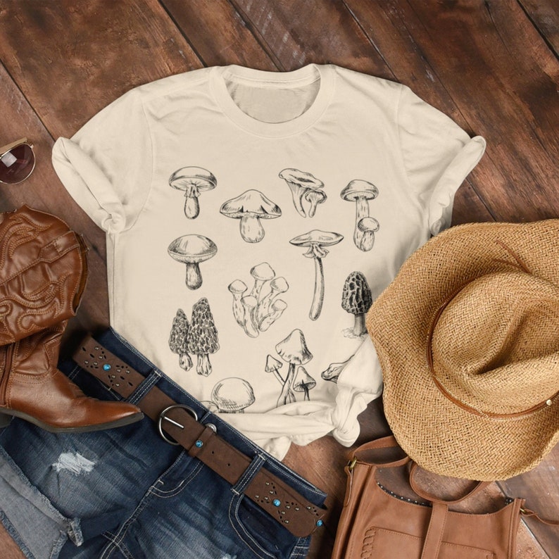 Puede incluir: Una camiseta beige con una ilustraci&oacute;n en blanco y negro de varios tipos de setas. La camiseta est&aacute; colocada sobre una superficie de madera con un par de botas de vaquero marrones, una bolsa de cuero marr&oacute;n, un sombrero de paja marr&oacute;n y un par de pantalones vaqueros azules con un cintur&oacute;n marr&oacute;n.