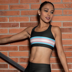 May include: A black sports bra with a transgender pride flag design. The bra has a racerback style and is made of a stretchy, breathable fabric.