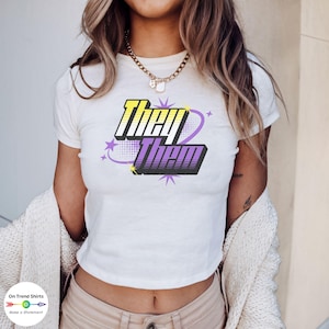 May include: A white crop top with short sleeves featuring a graphic design of the words "They Them" in a purple and yellow colour scheme with a starburst design.