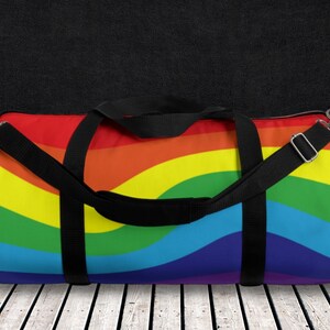 May include: A rainbow-coloured duffel bag with black straps and a mesh pocket. The bag has a rainbow pattern with red, orange, yellow, green, blue, indigo, and violet stripes.