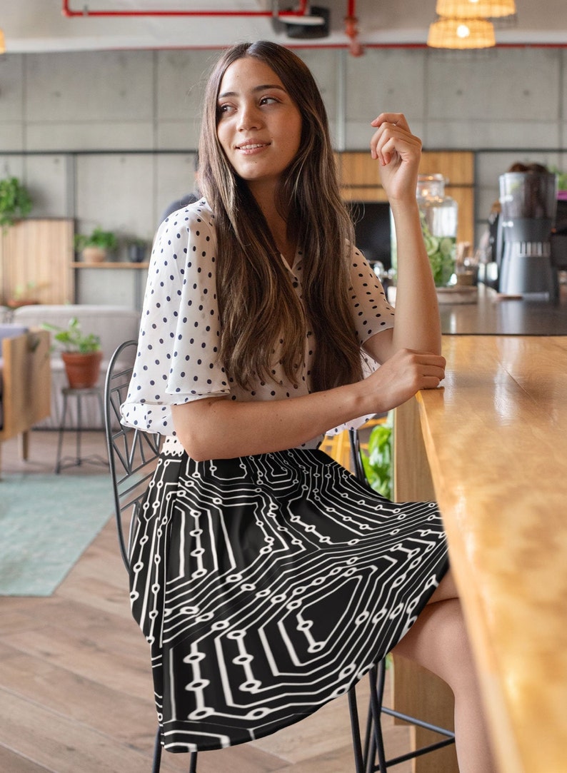 May include: A woman wearing a white polka dot blouse and a black skirt with a white circuit board pattern. She is sitting on a black metal chair in a cafe.