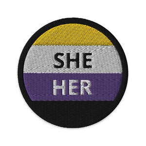 以下が含まれることがあります： 「SHE HER」という白い文字が、紫、白、黄色の背景に刺繍された丸いパッチ。パッチは黒い縁取りで囲まれている。