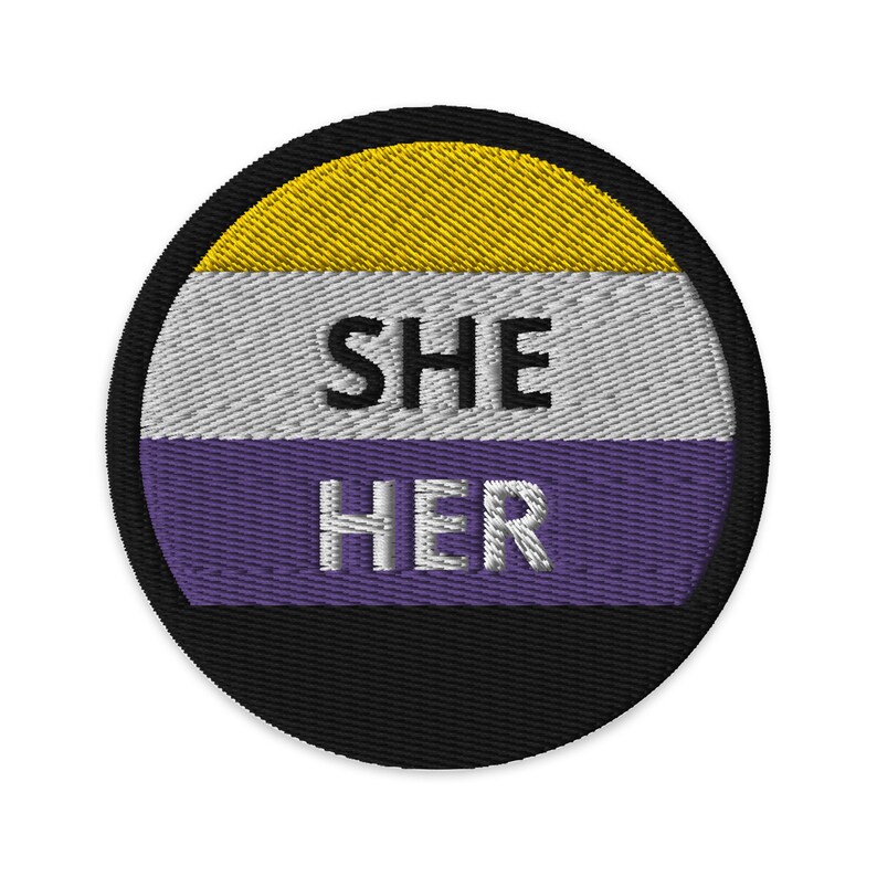 以下が含まれることがあります： 「SHE HER」という白い文字が、紫、白、黄色の背景に刺繍された丸いパッチ。パッチは黒い縁取りで囲まれている。