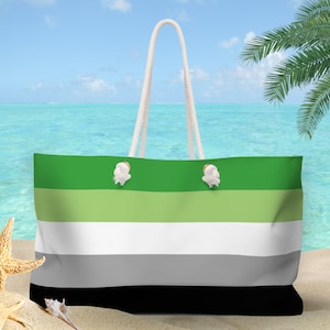 May include: A green, white, gray, and black striped tote bag with rope handles. The bag is on a sandy beach with a blue ocean and a palm tree in the background.