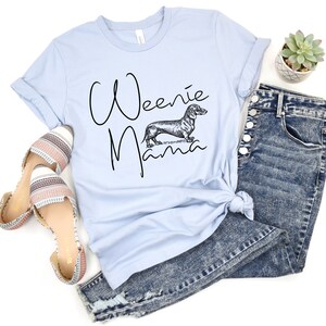 May include: Light blue t-shirt with the text "Weenie Mama" in black script and a black and white illustration of a dachshund. The shirt is paired with blue jeans and striped sandals.