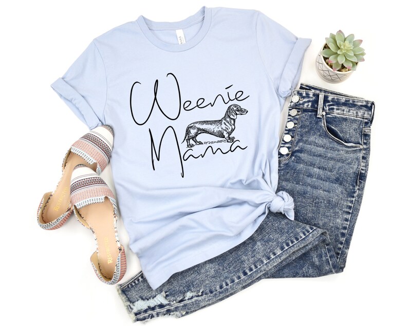 May include: Light blue t-shirt with the text "Weenie Mama" in black script and a black and white illustration of a dachshund. The shirt is paired with blue jeans and striped sandals.