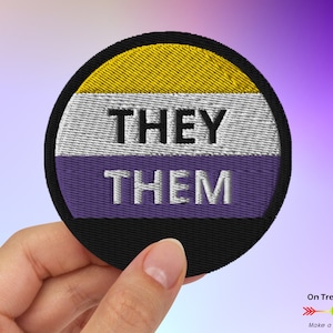 May include: A round embroidered patch with the text "THEY THEM" in white and black lettering on a purple, white, and yellow background. The patch is held in a hand.