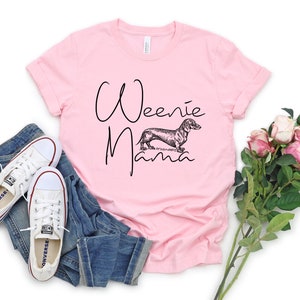 May include: Pink t-shirt with the text "Weenie Mama" in black script and a black and white illustration of a dachshund.