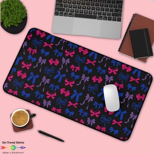 May include: Black rectangular mouse mat featuring a pattern of pink, blue, and purple bows. A white computer mouse is on the mat, with a laptop, notebook, and coffee cup in the background. The text "On Trend Shirts" is visible.