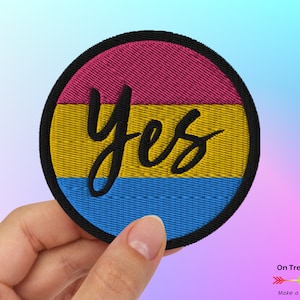 May include: A round embroidered patch with the pansexual pride flag colors: pink, yellow, and blue. The word "yes" is written in black cursive on the yellow stripe.