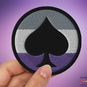 May include: A round, embroidered patch featuring a black spade in the centre. The spade is surrounded by three horizontal stripes: grey, white, and purple. The patch is held in a hand.