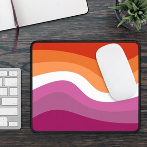May include: A lesbian pride flag mousepad with a white computer mouse on top. The mousepad features a wavy design with orange, pink, and white stripes.