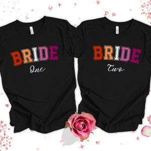 Puede incluir: Dos camisetas negras con la palabra "BRIDE" en colores arcoíris. Una camiseta dice "One" y la otra dice "Two".